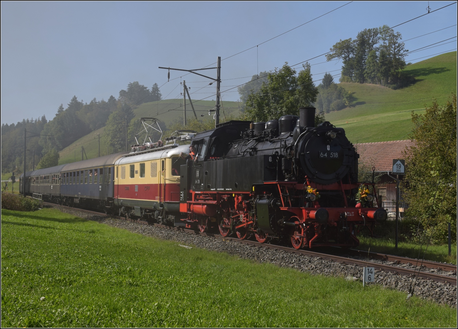 Dampftage Huttwil mit frisch genesenem Bubikopf. 

64 518 und Re 4/4 I 10034 mit zwei Silberlingen und einem erstklassigen Schnellzugwagen der DB kommt in Dürrenroth an. Rauch und Dunst stammen aber nicht von ihr selbst, sondern die im Bahnhof wartende Ec 3/4 2 der Solothurn-Münster-Bahn sorgte als Nebelmaschine für Stimmung. Oktober 2023