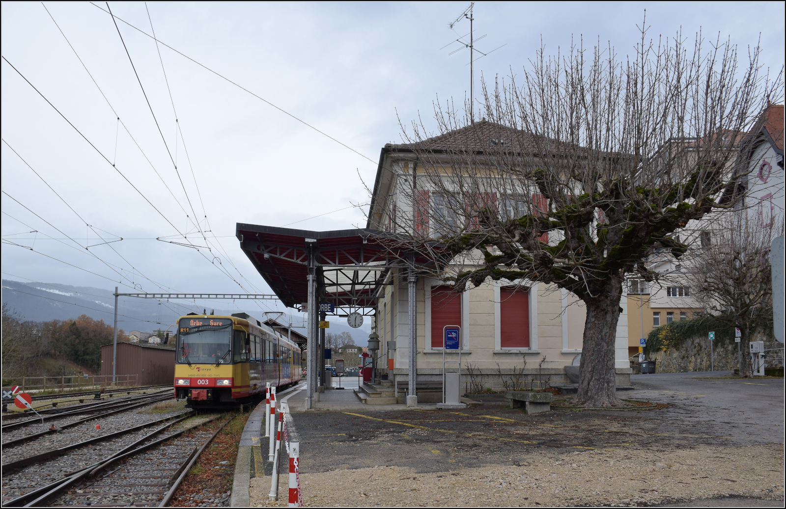 Ende einer Ära oder Gleichstrombetrieb adieu.

Die Strassenbahn  Be 4/8 003  alias 94 800 450 003-9 D-TVYS wartet in Orbe auf die nächste Ausfahrt nach Chavornay, um die Fahrgäste von den S-Bahnen dort abzuholen. Man beachte auch ein im Bild winziges Detail, das Läutewerk ist noch am Empfangsgebäude installiert. Dezember 2025.